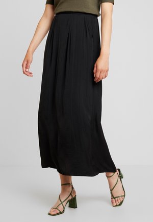 Femme portant une jupe midi plissée noire, un haut vert olive et des sandales vertes à talons avec brides, debout sur un fond uni.
