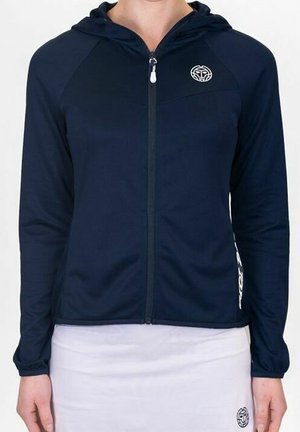 Kobieta ubrana w granatową bluzę z kapturem zapinaną na zamek, z białym okrągłym logo, oraz białą spódnicę z pasującym logo.