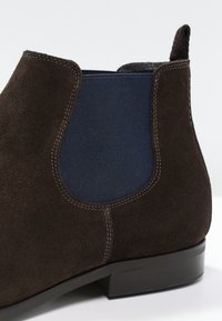 Bottine en daim marron avec un panneau élastique bleu, un bout arrondi et un petit talon en caoutchouc noir. Des détails de couture soignés sont visibles.