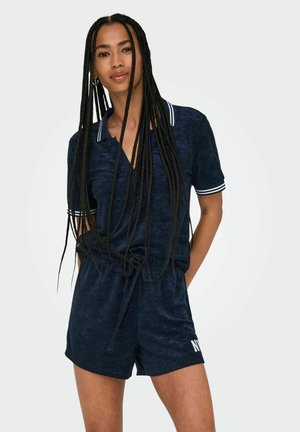 Marineblauer kurzer Overall aus weichem Material mit V-Ausschnitt und kurzen Ärmeln, mit weißen Akzenten am Kragen und an den Hosen. Verstellbare Taille.