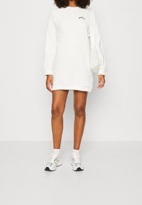 Vestido sudadera blanco con mangas largas, escote redondo y pequeño detalle de texto "WORLD". Combinado con zapatillas deportivas blancas y grises.