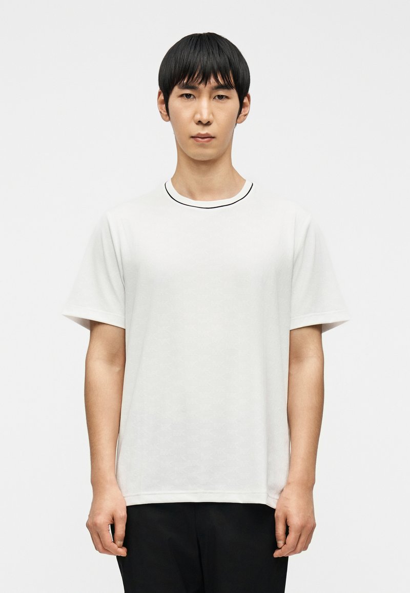 Jeune homme aux cheveux noirs et raides, portant un t-shirt blanc uni à manches courtes et un pantalon noir, debout devant un fond blanc.