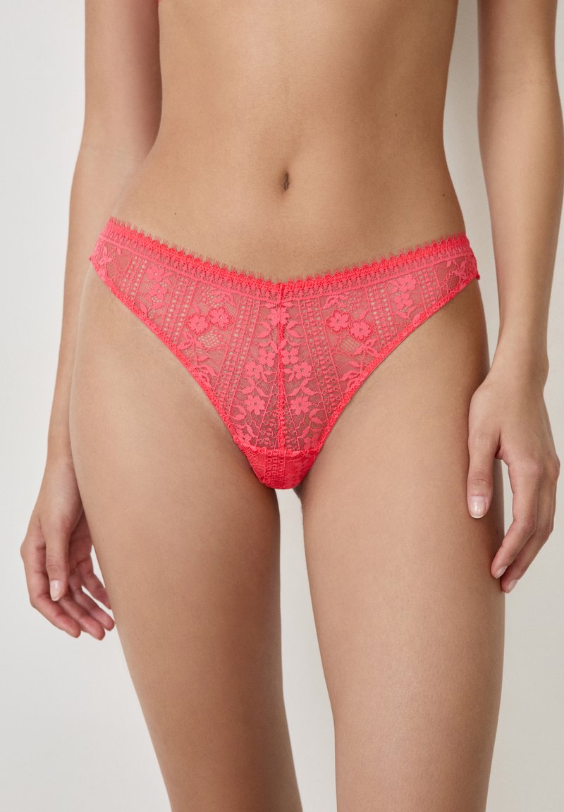 Spitzenstring in leuchtendem Pink, mit floralem Muster, filigranen Details und einem gewellten Bund. Weicher, transparenter Stoff überall.