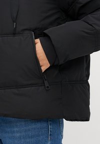 Mano parcialmente dentro del bolsillo con cremallera de una chaqueta acolchada negra sobre unos vaqueros azules.