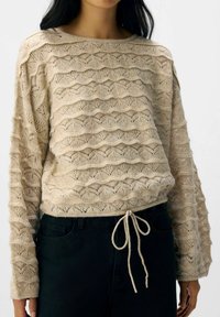 Beige mönstrad stickad tröja med vågiga horisontella texturerade ränder och dragsko i fållen, bärs med svarta jeans.