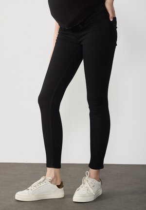 Person wearing black slim-fit pants and white low-top sneakers standing on a gray floor against a white background.