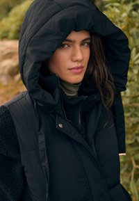 Gilet en duvet noir avec une grande capuche, comportant une fermeture éclair et des boutons-pression. Superposé à un vêtement sombre et texturé.