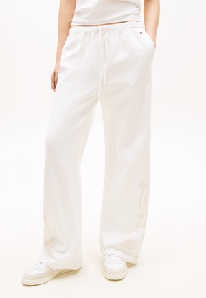 Άτομο που φοράει ευρύχωρο άσπρο παντελόνι φόρμας με διακριτικά κεντημένα μοτίβα κοντά στους αστραγάλους, συνδυασμένο με άσπρα αθλητικά παπούτσια και άσπρο τοπ.