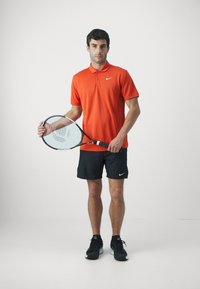 Rød polo skjorte med krage, svarte shorts, holder en tennisracket, og har på seg svarte sko med lyse detaljer. Minimalistisk design og merking.