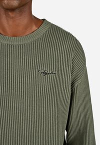 Zielony sweter prążkowany z okrągłym dekoltem, z czarnym haftowanym logo na lewej piersi. Teksturowany materiał i dopasowany krój.