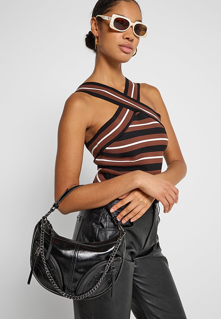 Schwarze lackierte Schultertasche mit Kettenriemen, mit geschwungenen Nähten und Reißverschlussdetails. Kombiniert mit einem gestreiften Oberteil und schwarzen Hosen.