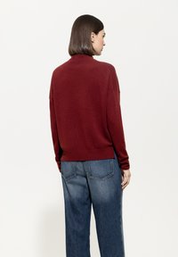 Frau mit schulterlangen braunen Haaren steht mit dem Rücken zur Kamera, trägt einen roten Pullover und lockere blaue Jeans vor einem schlichten weißen Hintergrund.