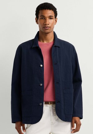 Jonge man met kort krullend haar, gekleed in een donkerblauw jasje over een rood overhemd en witte broek, staand tegen een effen lichte achtergrond.