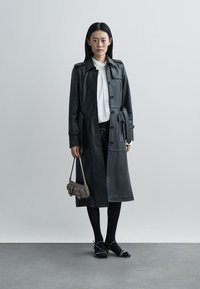 Długi czarny skórzany płaszcz trencz z zapięciem na guziki, bocznymi kieszeniami i paskiem. Połączony z białą koszulą, czarnymi spodniami i małą torebką.