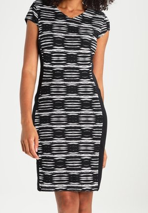 Femme portant une robe ajustée à motif noir et blanc avec des manches courtes et des panneaux latéraux noirs unis, debout devant un arrière-plan uni.