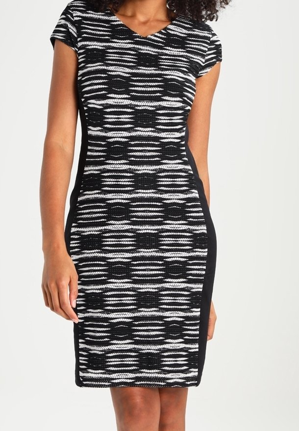 Femme portant une robe ajustée à motif noir et blanc avec des manches courtes et des panneaux latéraux noirs unis, debout devant un arrière-plan uni.