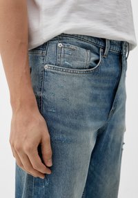 Jeans de mezclilla azul claro desgastados con detalles deshilachados, diseño de cinco bolsillos y acentos metálicos en la cinturilla y cierres de botones.