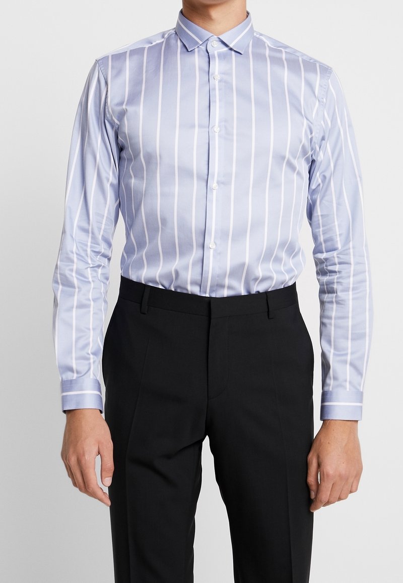 Muž oblečený ve fitted světle modré košili s bílými vertikálními pruhy a černými přizpůsobenými kalhotami, stojící na čistém pozadí.