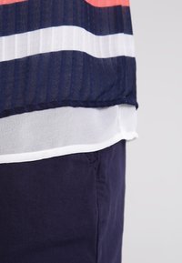 Top blu navy con strisce verticali bianche e corallo, con un orlo trasparente bianco. Tessuto morbido e leggero.