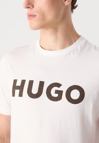 Fehér póló, amelynek középpontjában a merész barna "HUGO" felirat áll, herringbone mintázatban. Kerek nyakkivágású, rövid ujjú, sima textúrájú.