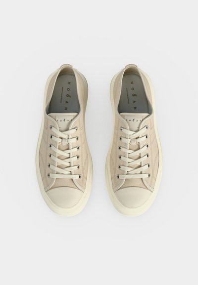Ζευγάρι μπεζ χαμηλά πάνινα παπούτσια με λευκές λαστιχένιες μύτες, μεταλλικά παπούτσια και κορδόνια, σε θέα από πάνω σε λευκό φόντο.
