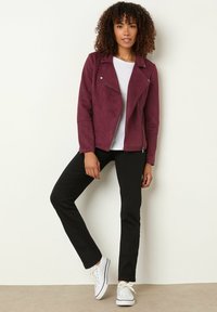 Veste en daim bordeaux avec fermeture éclair, design asymétrique, pantalon noir et baskets blanches. T-shirt blanc à col rond simple en dessous.