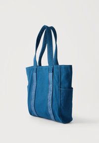 Μπλε τσάντα ώμου Calvin Klein με δύο λουριά ώμου και πλαϊνή τσέπη, σε όρθια θέση σε λευκό φόντο.