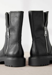 Bottes noires en cuir jusqu'aux chevilles avec surface texturée, fermetures éclair en argent et semelles en caoutchouc noir robuste. Dotées d'un talon épais et de coutures visibles.