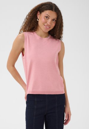 Jeune femme aux cheveux bouclés portant un haut rose sans manches et un jean en denim foncé, souriante devant un fond clair uni.