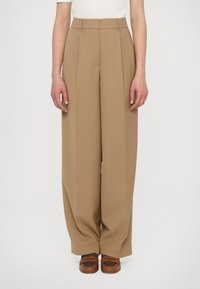 Pantalons taille haute plissés en tissu marron clair, avec une coupe évasée et une texture lisse. Assortis à des chaussures marron.
