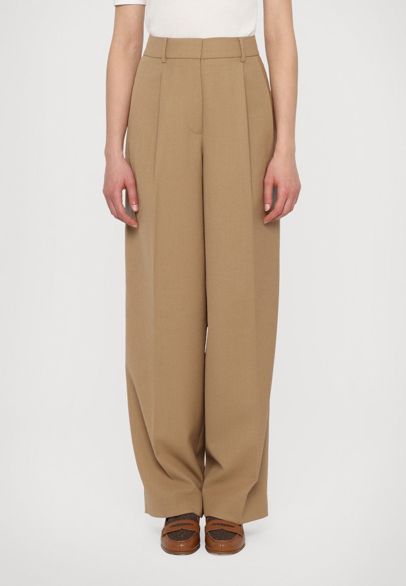 Pantalons taille haute plissés en tissu marron clair, avec une coupe évasée et une texture lisse. Assortis à des chaussures marron.