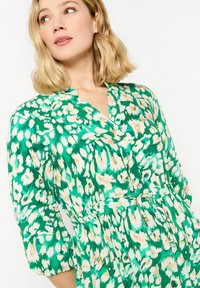 Abito verde con motivo floreale, scollatura a V, chiusura con bottoni e maniche a tre quarti. Tessuto morbido, vita arricciata e texture sottile.