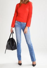 Röd långärmad topp med volangdetaljer, kombinerad med ljusblå jeans och svarta spetsiga klackskor. Håller en svart bucket-väska.