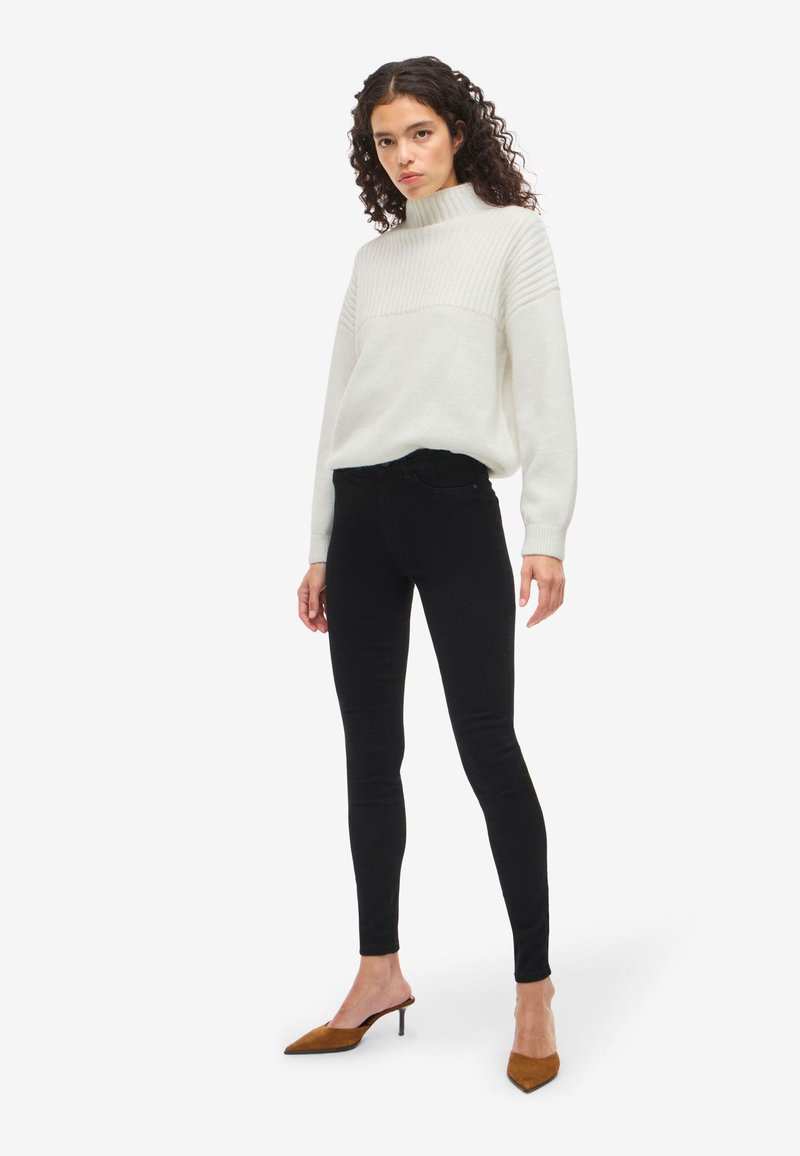 Bela pletena jopica z rebrastimi detajli in visokim ovratnikom, v kombinaciji s črnimi oprijetimi kavbojkami in rjavimi natikači s peto. Minimalističen dizajn.