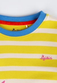 Gelb-weißer gestreifter T-Shirt mit einem hellblauen Kragen. Verfügt über ein pinkfarbenes Logo auf der linken Brustseite. Weicher Baumwollstoff.