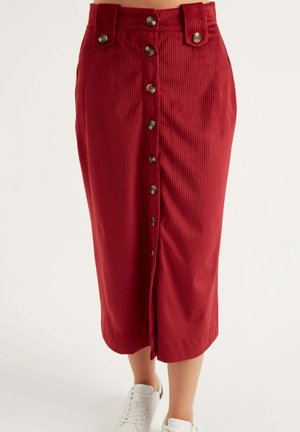 Jupe midi en velours côtelé rouge taille haute avec boutons sur le devant, passants pour ceinture, portée avec des baskets blanches, vue de la mi-cuisse jusqu'aux pieds.