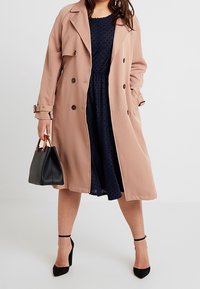 Ljusrosa trenchcoat med dubbelknäppt design, bärs över en marinvågsprydd klänning. Svart handväska och högklackade skor fulländar looken.