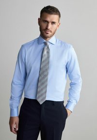 Chemise habillée à manches longues de couleur bleu clair avec un col pointu, associée à une cravate grise à rayures diagonales rouges et bleu marine, et un pantalon sombre.