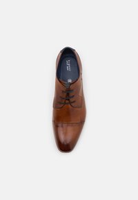 Chaussure de ville pour homme en cuir marron avec lacets, bout capé et panneaux latéraux texturés, vue de dessus sur un fond gris clair.