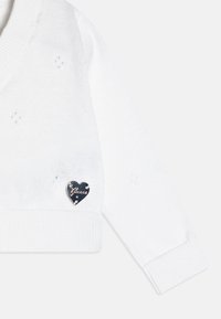 Balts adīts džemperis ar šalītu apkakli, kurā ir sirds formas melns logs akcenta kreisajā pusē un strukturēts raksts visā virsmā.