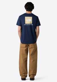 Marineblå bomulds t-shirt med et firkantet logo i cremede og hvide farver på ryggen, med teksten "NEVER STOP EXPLORING" under logoet.