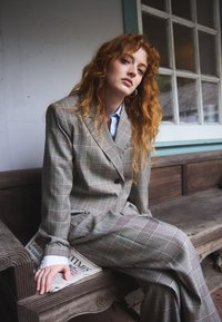 Femme aux cheveux roux en costume à carreaux gris assise sur un banc en bois tenant un journal plié près d'une fenêtre à plusieurs vitres.