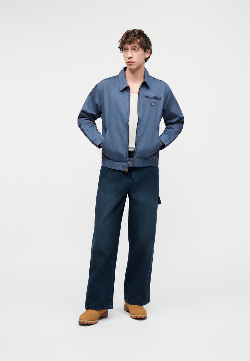 Jonge man met doorzichtige bril, blauw vest met ritssluiting, wit overhemd, brede donkere spijkerbroek en beige laarzen, staand met handen in de zakken.