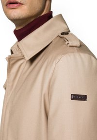 Beżowy trencz z wyraźnym kołnierzem i pagonami. Posiada teksturowany materiał, ciemną metkę na rękawie oraz bordowy golf pod spodem.