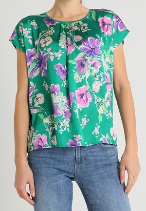 Zielona kwiecista bluzka z krótkimi rękawami, ozdobiona fioletowymi kwiatami, o luźnym kroju i delikatnych zakładkach przy dekolcie. Noszona z niebieskimi dżinsami.