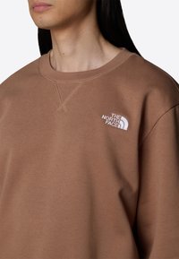 Sudadera marrón hecha de tela suave, con cuello redondo y logotipo blanco bordado en el pecho. El diseño tiene un ajuste holgado.