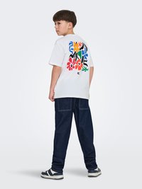 Jongen met een wit T-shirt met een kleurrijk abstract bloemmotief op de achterkant, donkerblauwe spijkerbroek en sneakers, staand en naar de zijkant kijkend.