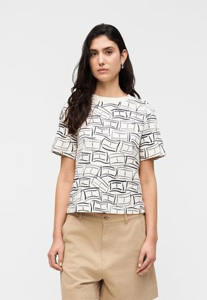 Femeie cu păr lung și închis la culoare poartă un tricou alb cu mâneci scurte cu modele abstracte negre și pantaloni scurți bej cu talie înaltă, stând neutră.