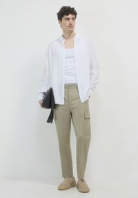 Camicia bianca a maniche lunghe, canotta semplice, pantaloni cargo kaki con tasche, scarpe slip-on color beige, tiene in mano una pochette nera a forma triangolare. Sfondo neutro.