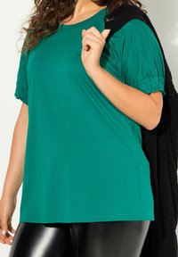 Top verde a maniche corte con maniche a sbuffo arricciate, realizzato in tessuto morbido. Abbinato a un maglione nero di tessuto con texture appoggiato sulla spalla.